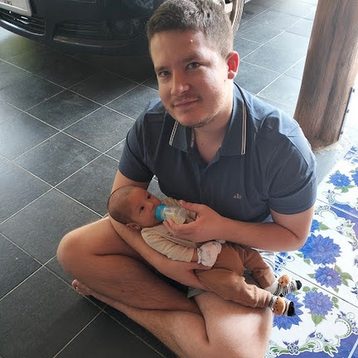 A young man feeding a baby with a bottle, sitting on the floor at home, showcasing family bonding and care.